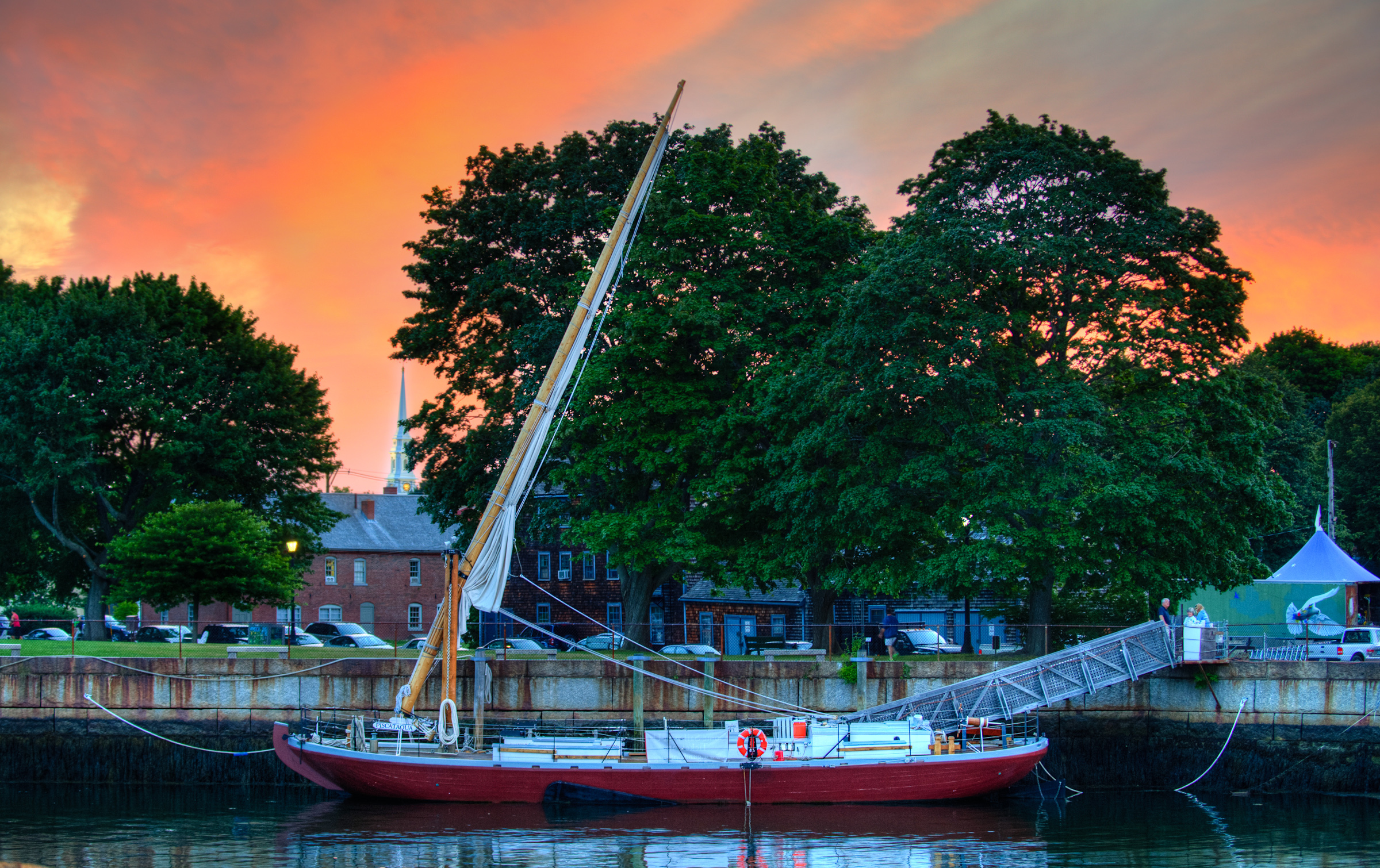 The gundalow Piscataqua sits docked in front of Prescott Park in Portsmouth NH at sunset. The steeple of the North Church is illuminated in the background by lights that have just come on.