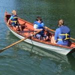 Two campers and one counselor row a white wherry with orange interior (a wherry is a historical type of wooden rowboat).