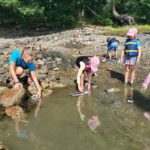 A counselor in training and a group of campers crouch at the end of a river shore to float and test their carboard and aluminum boats.