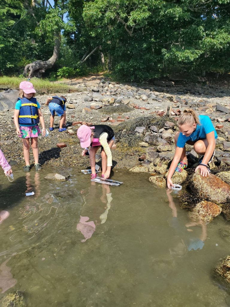 A counselor in training and a group of campers crouch at the end of a river shore to float and test their carboard and aluminum boats.